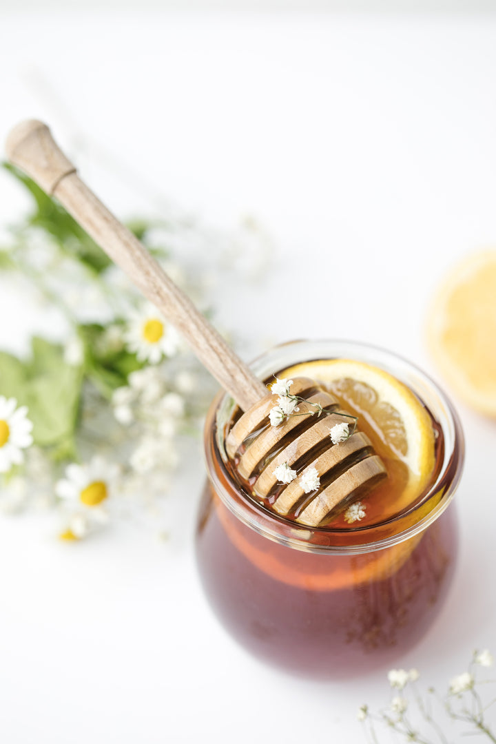 Wooden honey stick in jar of honey and lemon in front of flowers with text, “Sarrasin Honey: The new Superfood Ingredient For Healthier Skin”.