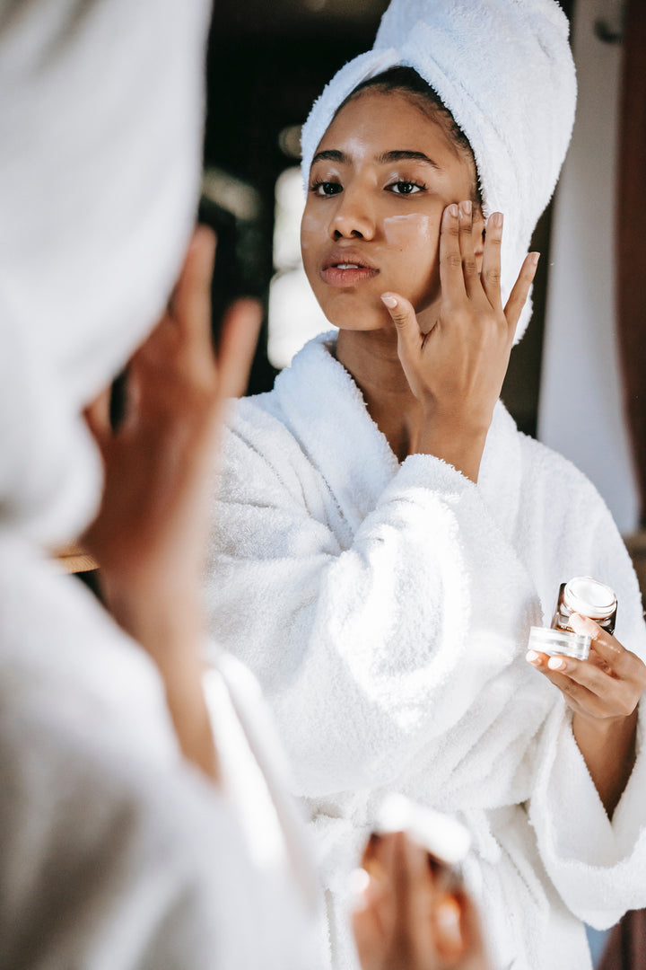 Woman in robe and hair towel applying face cream looking in the mirror.